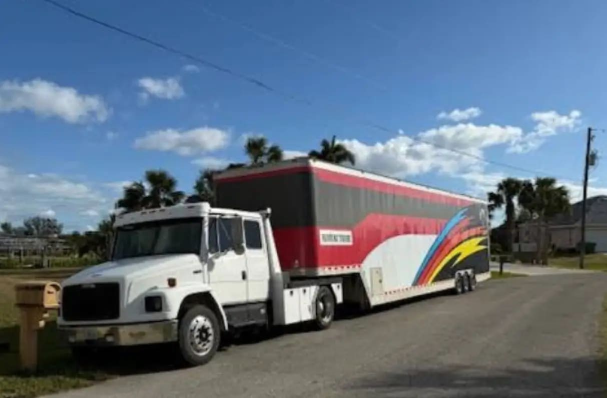 A white Freightliner tractor pulling a long enclosed car trailer along a residential street.