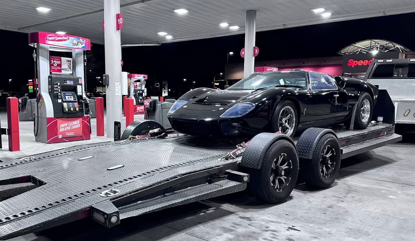 A black GT40 secured on a tilt-deck trailer under the canopy lights of a fuel station.