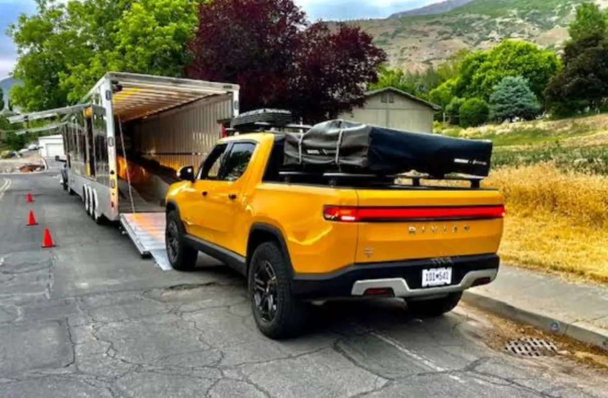 A yellow Rivian pickup with a rooftop tent being driven up the ramp into an enclosed trailer, traffic cones set out behind it.